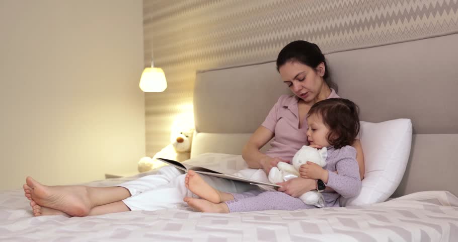 Mother reading fairytale to her little child daughter in bedroom. Education for children.