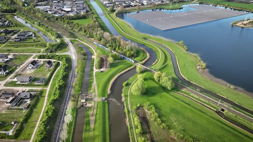 Aerial footage of floating solar farm on water, surrounded by canals and green spaces with a train crossing the landscape, residential areas and industrial zone. Sustainable infrastructure with urban.
