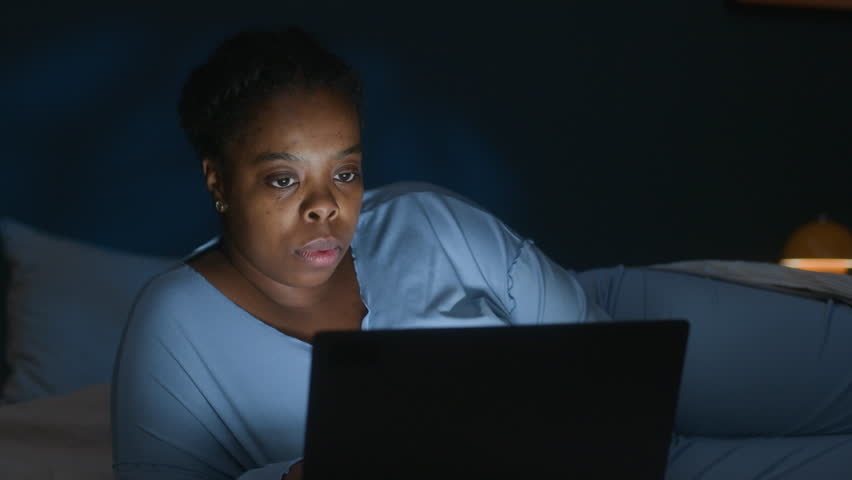 Young Black woman in blue pajamas resting in dark bedroom, typing on laptop during sleepless night at home