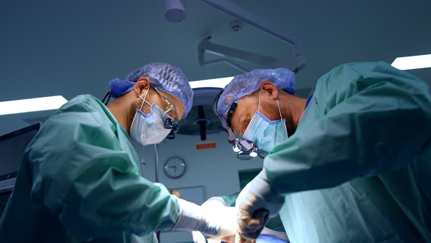 Two surgeon co-work at maxillofacial surgery. Attentive doctors in surgical loupes carefully use the instruments. Low angle view.