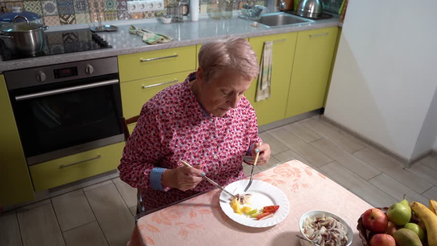 An elderly woman eats fish and vegetables on a plate with a knife and fork. A grandmother is having lunch at home in the kitchen. Healthy eating. Table top view