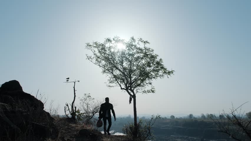 A steady wide silhouette shot of a Indian Biker man walking in his gear on a rocky path with some greenery and trees around on a sunny afternoon.