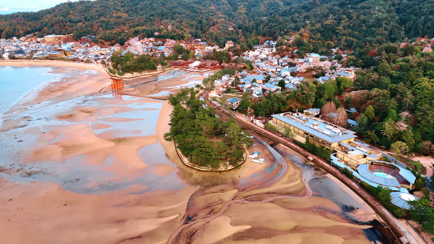 Multiple lovely houses surround the territory of sacred Itsukushima Shrine at the Miyajima island, Japan. Period of low tide at the sandy shore of the Inland Sea. Aerial view.