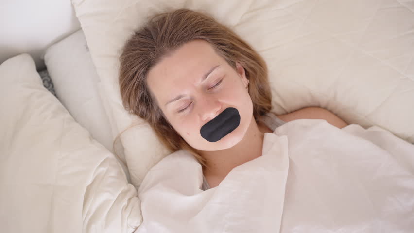 Woman Lying Peacefully in Bright Bedroom with Mouth Taped Shut Under White Blanket in Morning Light for Sleep Recovery Concept
