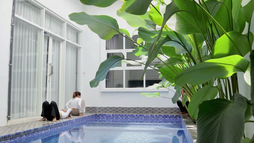 Two beautiful women in sportswear are near the aesthetic home pool doing yoga together. Meditation in a cool and peaceful area of ​​the house. Photographed by a photographer from behind the leaves.