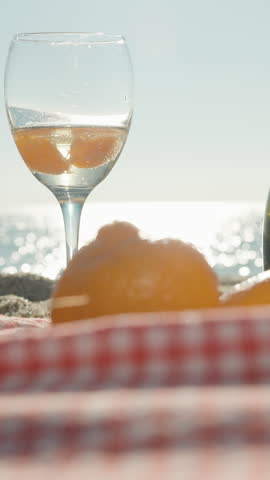 Vertical video. A Tangerine Slice Tossed Into a Champagne Glass on a Red Checkered Picnic Blanket with Oranges and a Champagne Bottle by the Ocean under Bright Sunlight