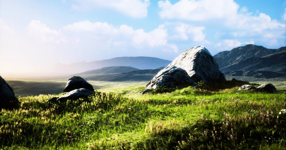 A serene landscape showcasing large rocky formations surrounded by lush green grass under a clear blue sky, with rolling hills in the background illuminated by the warm sunset light.