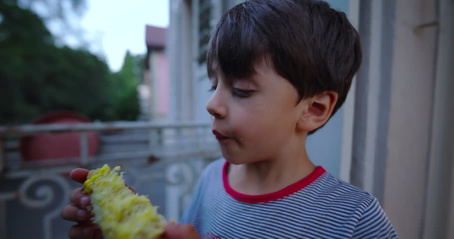 Side view of a boy enjoying corn on the cob, with corn kernels on his face, outdoors on a balcony during a quiet evening in a residential area