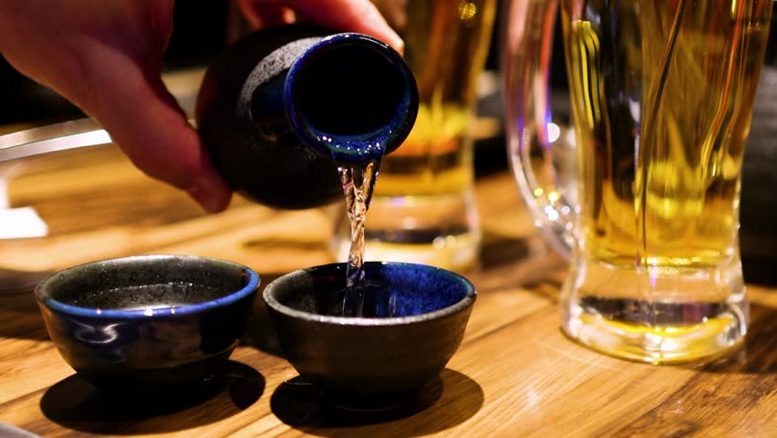 A hand pours sake into two blue ceramic cups beside tall beer glasses on a wooden table.