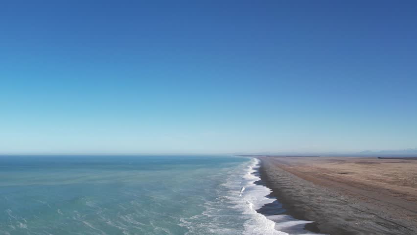 High aerial above incoming waves from beautiful turquoise ocean with flat, dry land and foothills in distance (Birdlings Flat Beach and Kaitorete Spit, New Zealand)