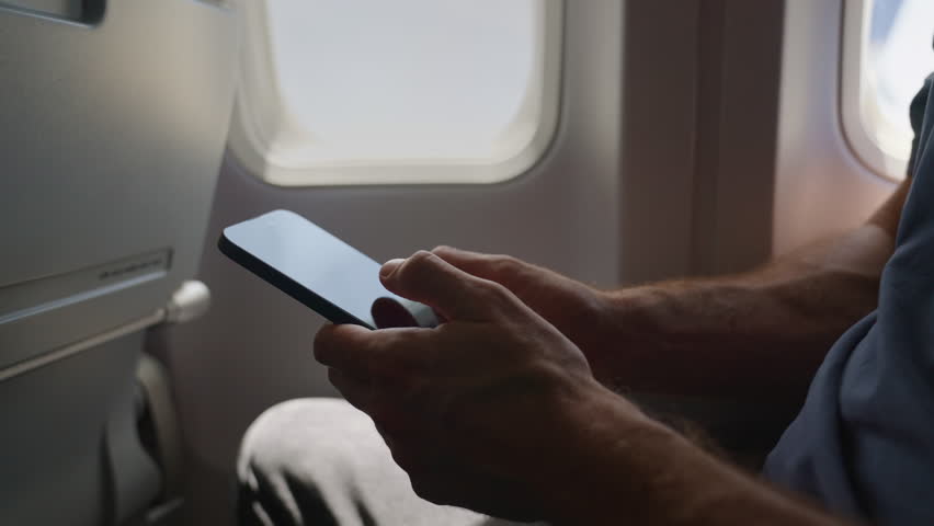 Wifi connection on flight. Close up on hands of male holding mobile phone and typing messages or using internet. Window seat. Modern airplane with special connection to use onboard aircraft