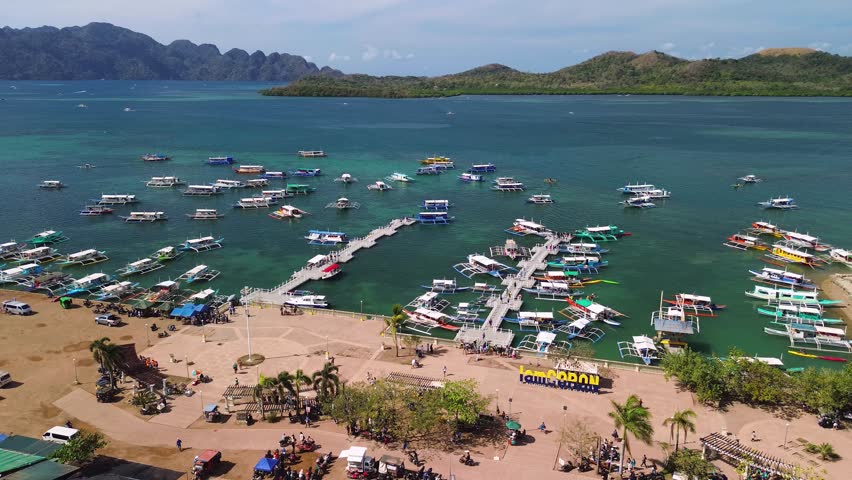 Drone footage of the main port in Coron, Palawan, where tourists begin their boat tours. Dozens of colorful bangkas docked at floating piers, with a clear sea and tropical mountains in view.