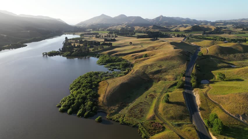 Aerial drone shot of golden hills, calm river, and winding road in morning light near the top of New Zealand’s South Island. Peaceful rural landscape with soft shadows and clear skies.