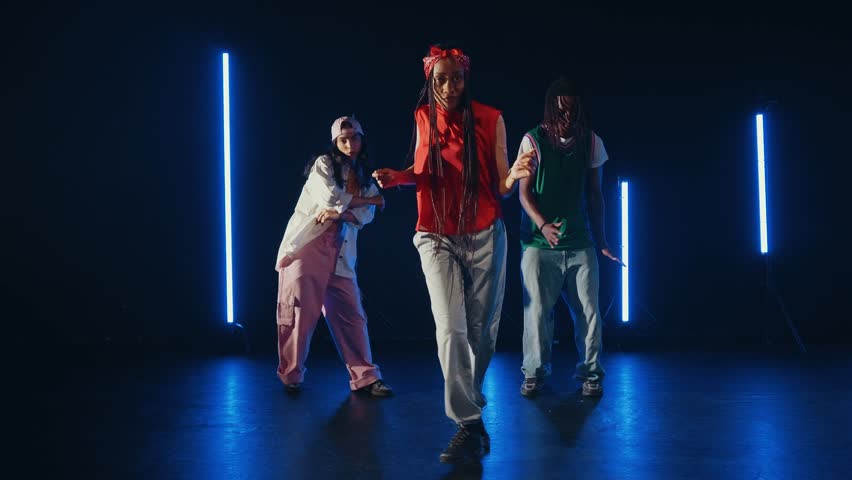Three talented dancers, two women and a man, showcase their impressive moves on a dark stage illuminated by vertical blue neon lights, creating a vibrant and dynamic performance