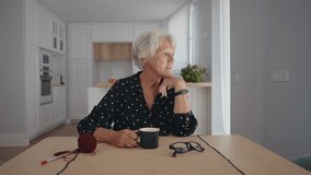 Elderly woman sits thoughtfully at her kitchen table, savoring a cup of coffee while gazing out the window, with knitting supplies nearby suggesting a peaceful morning routine - Powered by Shutterstock - Get 15% off with code: PIKWIZARD15