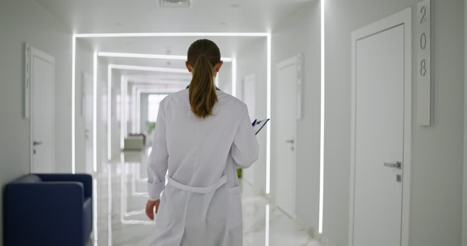 Rear view of female doctor in white coat holding tablet in hands walking down corridor and entering needed office during work in hospital