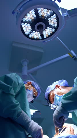 Caucasian surgeons cooperate at operation. Doctors use modern medical tools. Low angle view. Vertical video.