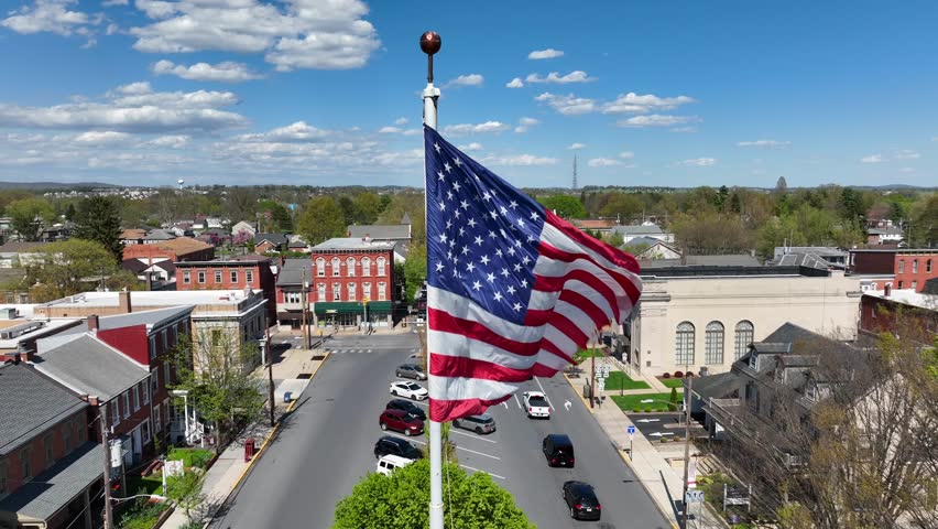 Patriotic American flag waves over small town in USA. Main Street in Small Town America square at sunny day in spring. Aerial close up orbit shot.