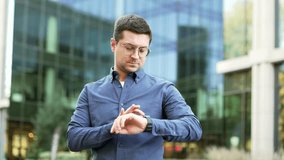 Confident businessman using smartwatch while standing on city street near modern office building. Business man with glasses looks at smart watch uses an app. Handsome male worker checks notifications - Powered by Shutterstock - Get 15% off with code: PIKWIZARD15