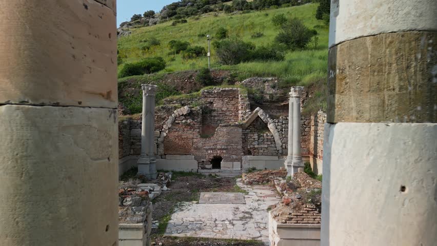 Aerial view of ancient Ephesus ruins with two tall stone columns. Green hills in background. Historic site in Turkey reveals remnants of classical architecture and Roman influence