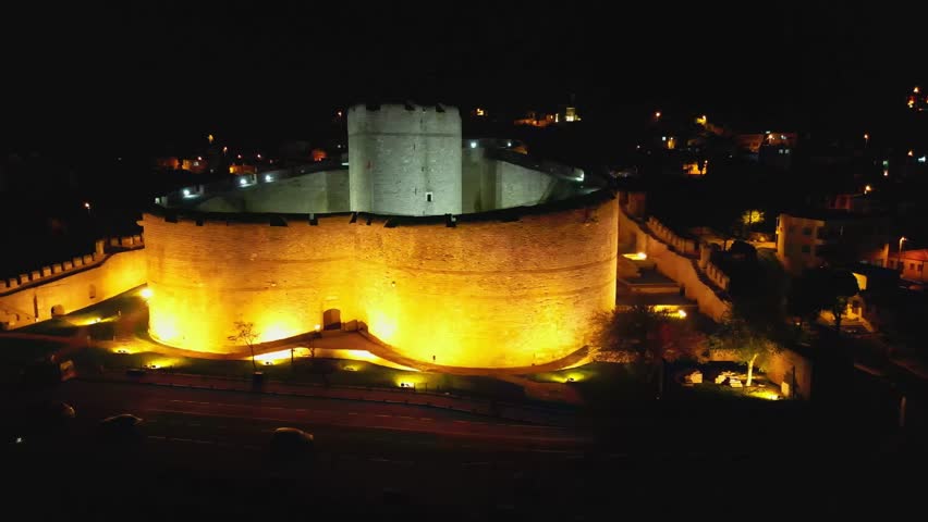 A fortress stands glowing in yellow lights along the dark coast. A side perspective reveals its massive round towers and strong walls, preserving Ottoman architectural heritage by the sea