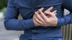 Close up. Male hands holding chest. Man in blue shirt has heart attack sitting on bench on street near business office building. Sick man massaging a painful sore spot, needs urgent medical attention - Powered by Shutterstock - Get 15% off with code: PIKWIZARD15