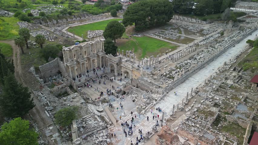 Oblique aerial angle shows Roman amphitheater and historic buildings in Ephesus. Tourists walk among ancient stones. Structure reveals grand design and rich cultural history of the region.