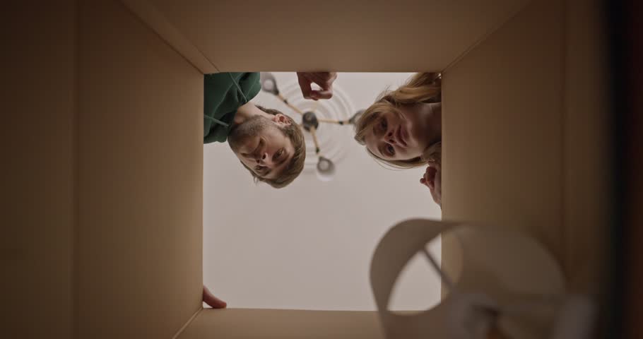 A guy and his blonde girlfriend take a table lamp from a large cardboard box while moving to a new apartment