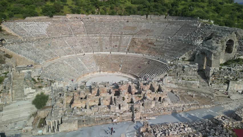 High angle view of the grand Roman amphitheater in Ephesus, Turkey. Ancient stone seating encircles a central stage. Visitors explore the vast historic ruins below green hillside