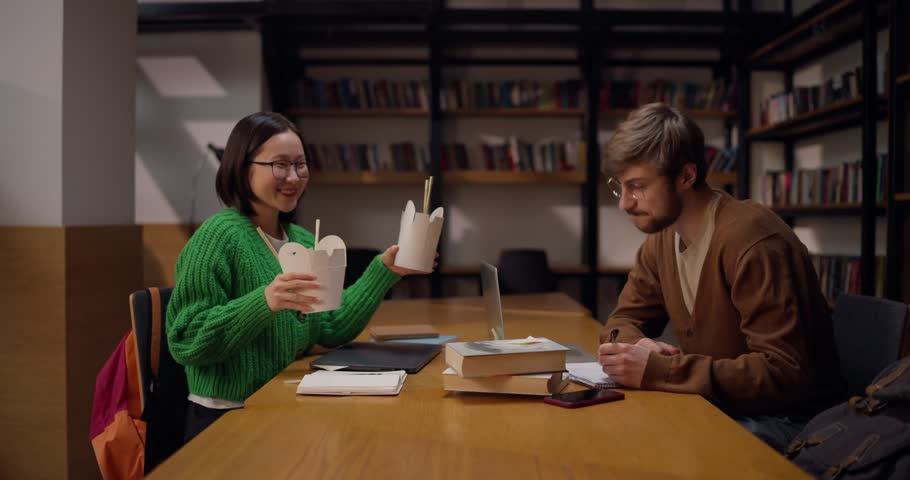 A girl with glasses hands her student colleague a lunch box with noodles during a break and lunch in the library