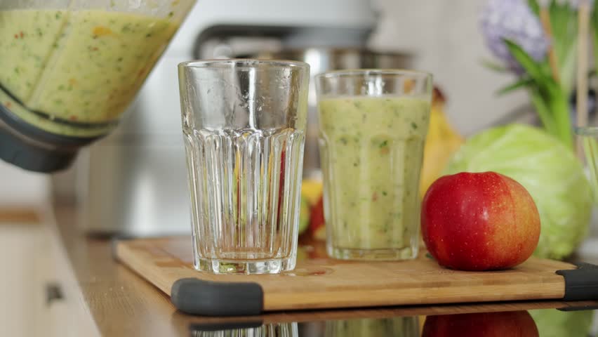 Female hands pouring fresh healthy green smoothie from apples, banana, broccoli, greens, celery from blender into glasses in kitchen at home. Detox breakfast drink to cleanse and provide energy body.