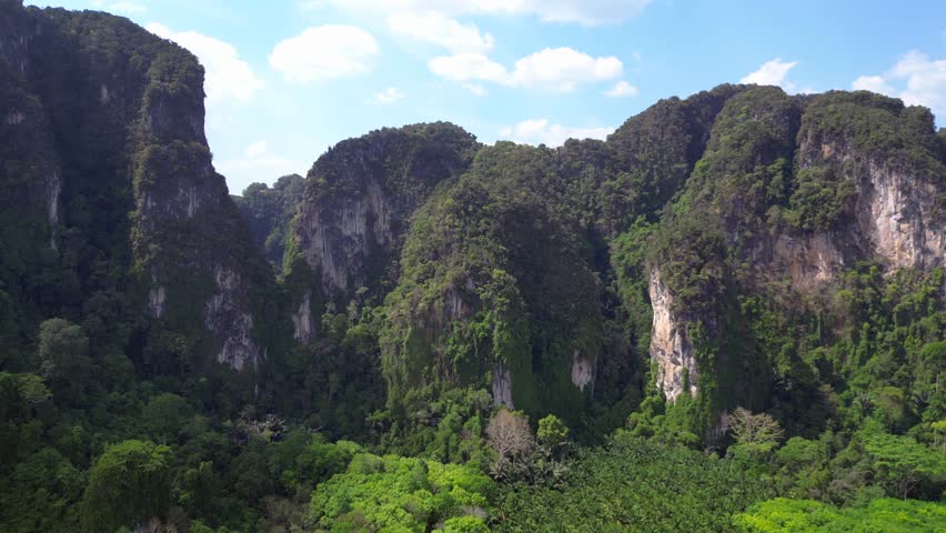 Lush green forest is covering the valley floor below towering limestone mountains in krabi, thailand. Amazing aerial view flight fly reverse drone
