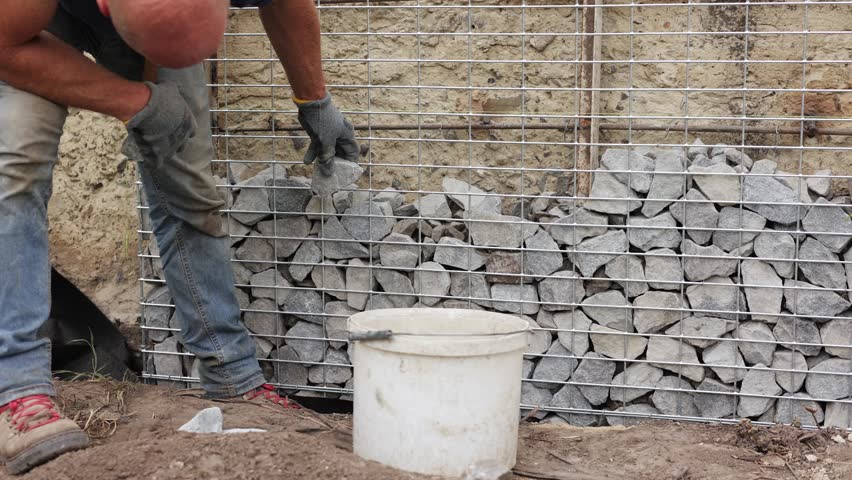 Stone fence in steel mesh. Close up of unrecognisable professional worker builds gabion and tightly packs stones of varying sizes and textures. Combination of natural materials and industrial design.