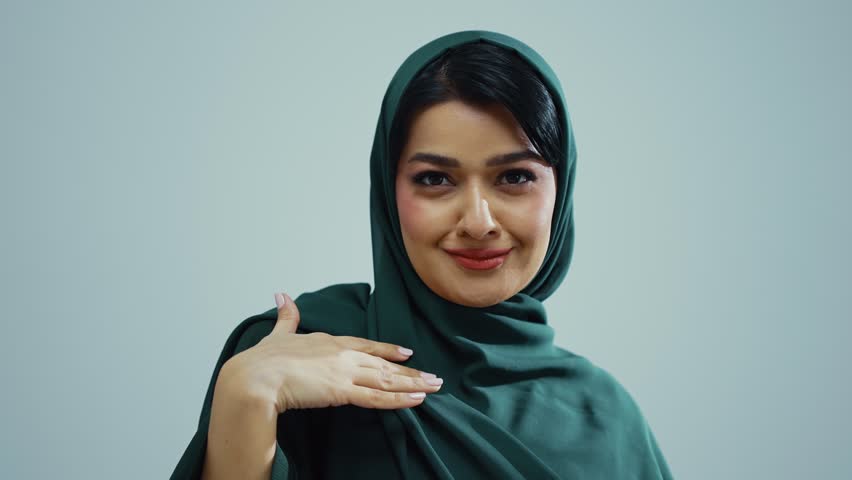 Studio portrait of an attractive young muslim woman wearing a traditional dark green hijab, posing on a white background, looking at camera and smiling.