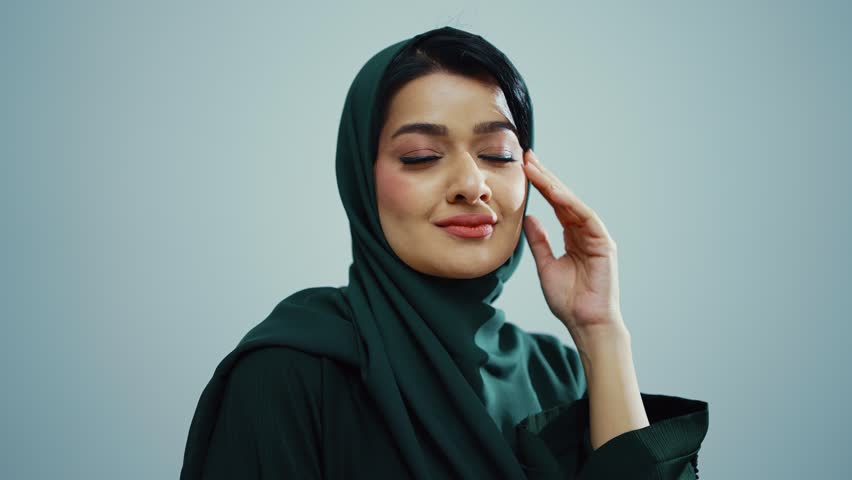 Studio portrait of an attractive young muslim woman wearing a traditional dark green hijab, posing on a white background, looking at camera and smiling.