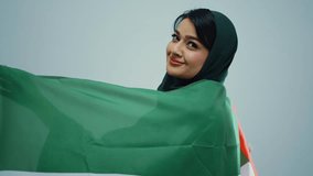 Studio portrait of an attractive young muslim woman wearing a traditional dark green hijab, posing on a white background, looking at camera and smiling while moving with uae flag. - Powered by Shutterstock - Get 15% off with code: PIKWIZARD15