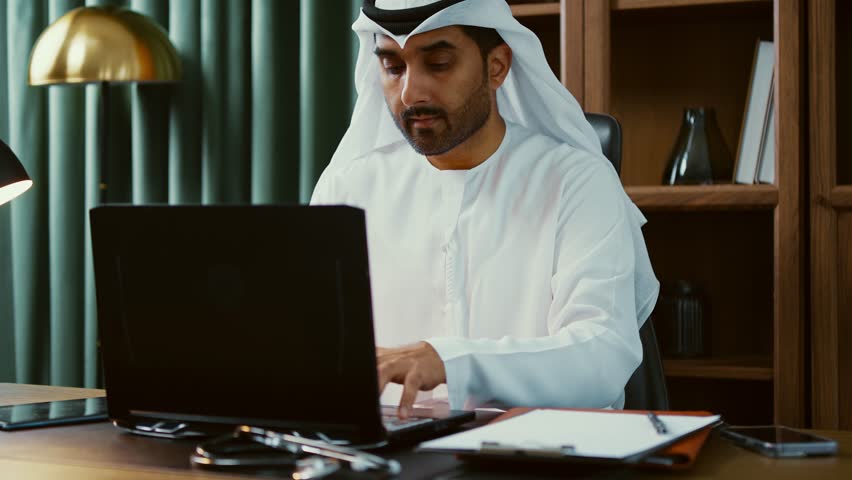 Professional emirati businessman wearing white kandura, working diligently on laptop in modern office, showcasing technological expertise within sleek corporate environment