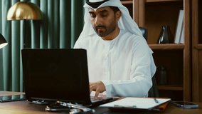 Professional emirati businessman wearing white kandura, working diligently on laptop in modern office, showcasing technological expertise within sleek corporate environment - Powered by Shutterstock - Get 15% off with code: PIKWIZARD15