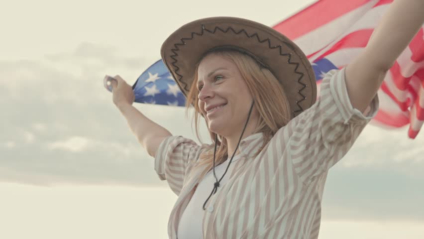 Blonde woman celebrating national pride, wearing cowboy hat, waving usa flag with wide-open arms against cloudy background.Usa celebrate 4th of july. Independence day concept