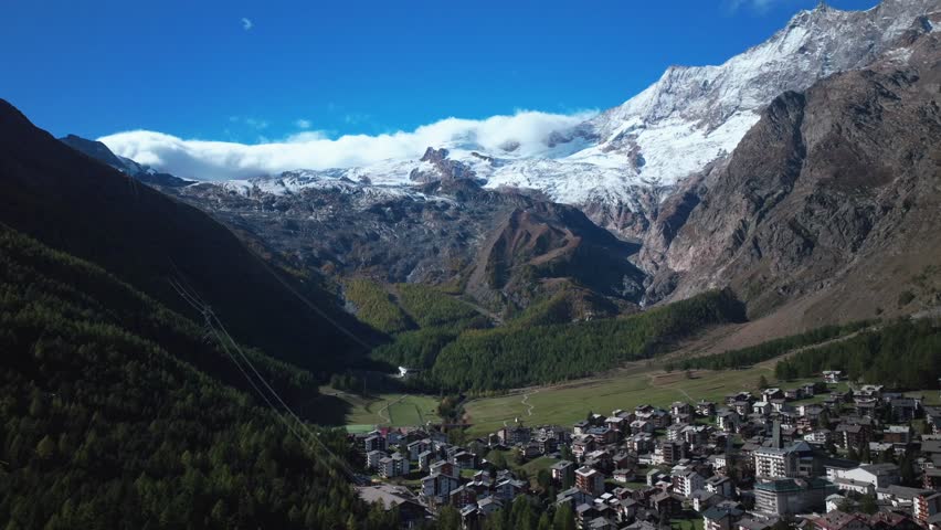Sunny stunning Saastal Saas Fee village town Switzerland aerial drone beautiful Fall Autumn Swiss Alps mountain peaks glacier surrounding city buildings bridge Zermatt The Matterhorn backwards