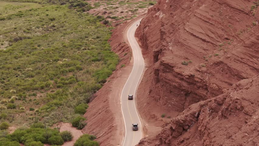 Aerial view of the road in Calchaquí Valley, with red rocks on the right and green wild grass on the left, while cars drive through the valley. Salta Argentina.