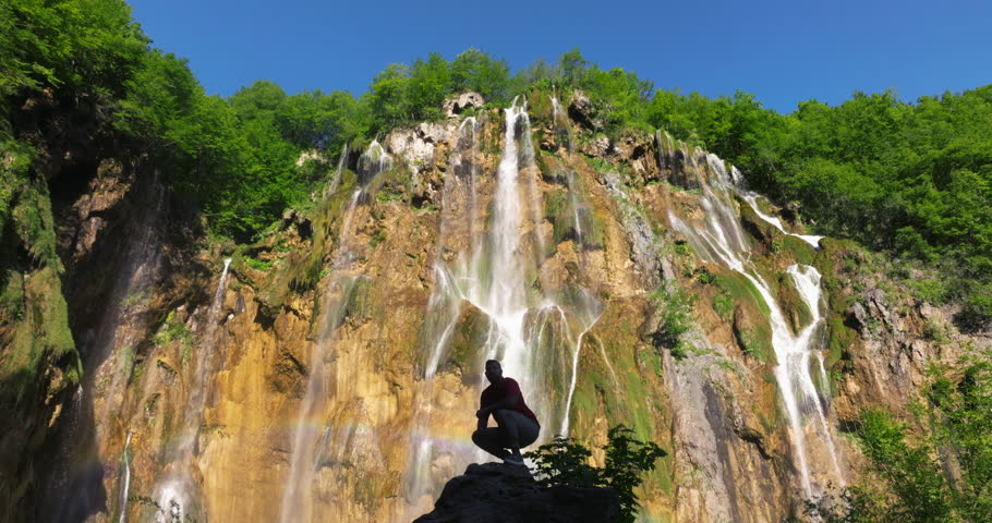 Male Tourist Stands On Rock Against Veliki Slap Waterfall In Plitvice Lakes National Park, Croatia. ascending drone shot