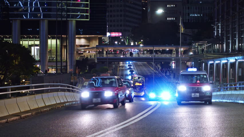 Nighttime cityscape of Hong Kong featuring busy streets filled with cars. The scene includes illuminated skyscrapers, traffic lights, and pedestrians. Dynamic urban environment
