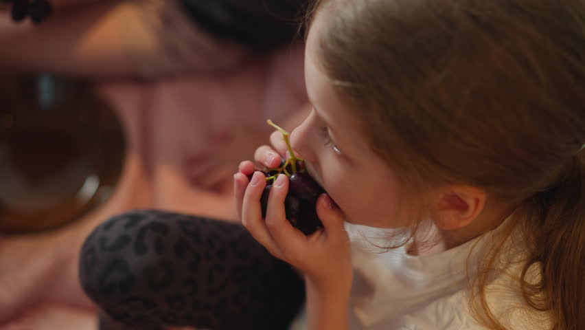Top view of little girl eating branch of grape holding tightly with tiny hands, soft natural light highlighting tender moment, blurred background