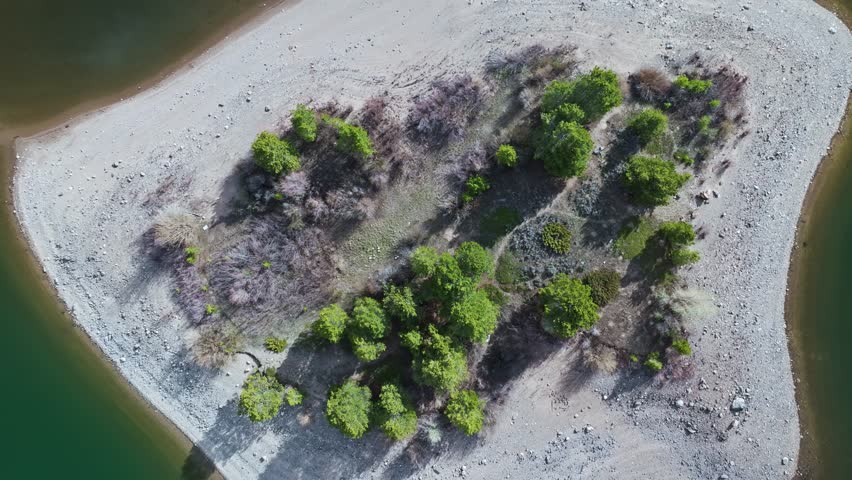 Drone rises above sandy island with evergreen trees casting longshadow across water