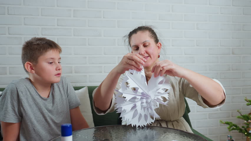 mother carefully unfolding handcrafted paper ornament while son watches closely with serious expression sitting together indoors at glass table against white brick wall decorated with simple cushions