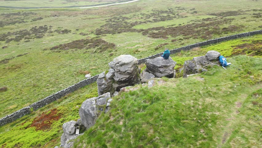 A phenomenal aerial shot over a solo traveller sitting on the edge of the rocky hill. A green trail is running along the hill