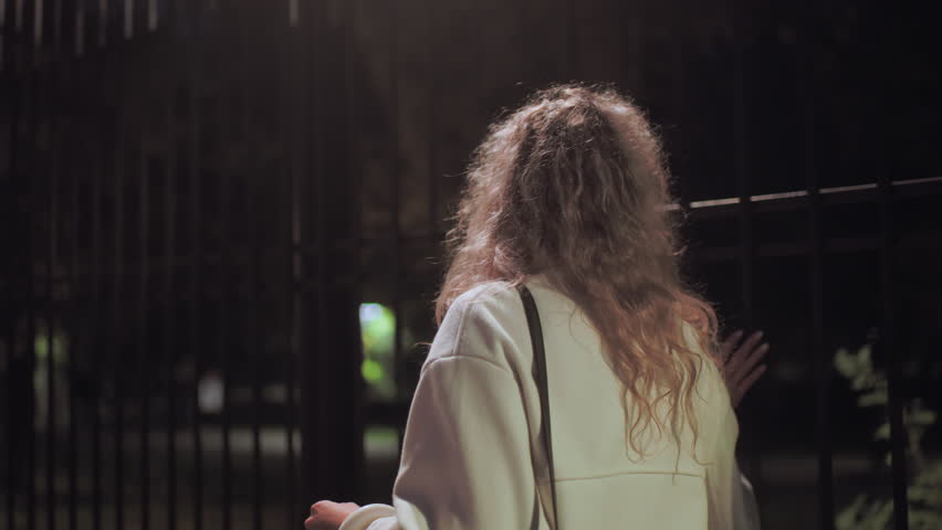 Woman in white coat walking with bag touching iron fence during peaceful night stroll with soft blurred background showing people walking down sidewalk and parked cars illuminated by streetlights