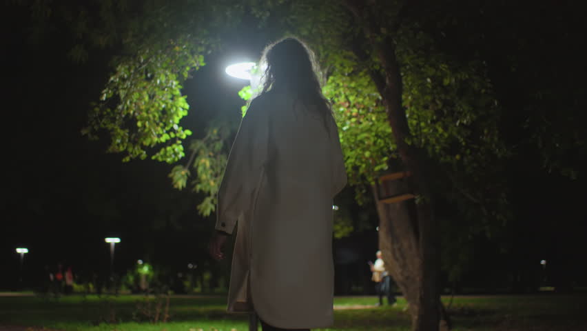 Back view of young white girl walking along dimly lit park path at night, holding umbrella, surrounded by glowing streetlights, with several blurred figures moving in distant background among trees