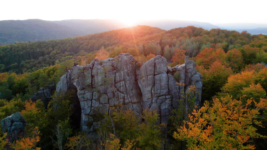 Aerial view of sun sets behind rugged rock formation, casting warm glow over landscape. Trees in autumn colors surround rocks, creating vivid contrast. Dovbush Rocks, Carpathian mountains, Ukraine.